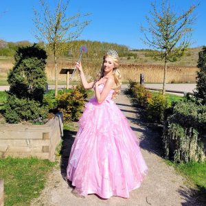 Picture showing a blonde good witch in a pink ballgown with trees behond her on a sunshiney day