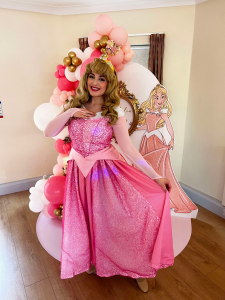 A female children's entertainer dressed as a classic fairytale princess in a pink gown, long blonde wig, and golden crown. She’s standing in front of a themed balloon display with pink, white, and gold balloons, and a cardboard cut-out of the same princess character. She is smiling and holding out the skirt of her dress.