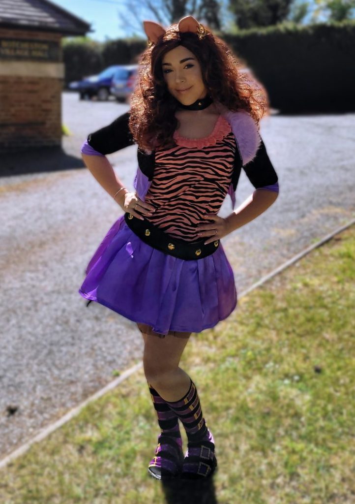 Person in a tiger-striped top, purple tutu skirt, cat ear headband, and striped socks posing outdoors on a sunny day.