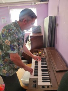 Man in floral shirt plays a brown organ with black and white keys.