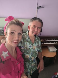 Two entertainers, one wearing a pink shirt with star embellishments and a pink headband, the other in a teal Stitch-patterned shirt, stand in front of a keyboard.