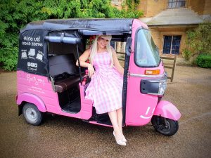 A person in a pink checkered dress sits joyfully in a pink tuk-tuk with