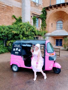 A woman dressed as Barbie in a pink plaid dress and white heels stands smiling beside a bright pink Rosie May tuk-tuk. The brick building behind her is partially covered in greenery, creating a lively and cheerful scene.
