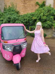 A woman dressed as Barbie in a pink checkered dress poses joyfully next to a pink tuk-tuk with