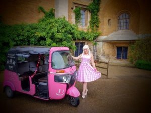 A woman dressed as Barbie in a pink gingham dress poses beside a pink tuk-tuk with a smiley face. The backdrop is a Belvoir Castle with ivy, creating a cheerful and whimsical scene.