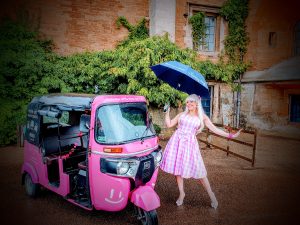 A woman in a pink plaid dress holds a blue umbrella, posing cheerfully beside a vibrant pink tuk-tuk. The background features ivy-covered brick walls.