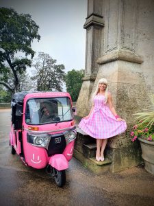 Woman in a pink checkered dress smiles beside a pink tuk-tuk with a happy face design. The scene is joyful, set near a stone column and trees.