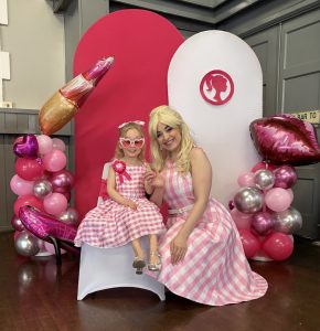 Barbie party entertainer with a child at a Barbie-themed birthday party, featuring pink balloon displays and themed seating.