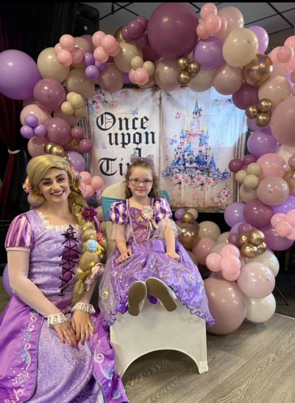 Princess entertainer dressed as Rapunzel posing with a child in a purple princess dress at a birthday party in Nottingham with pastel balloon backdrop and princess party setup by Absolutely Amazing Parties.