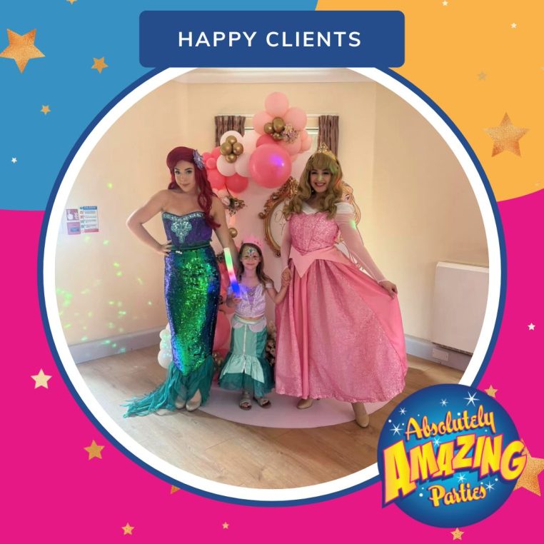 Two women in mermaid and princess costumes stand with a young girl in a matching outfit. They're smiling, in front of pink and gold balloons, under a 