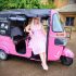 A person in a pink checkered dress sits joyfully in a pink tuk-tuk with