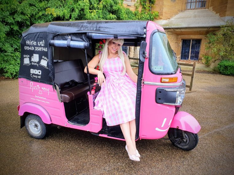 A person in a pink checkered dress sits joyfully in a pink tuk-tuk with
