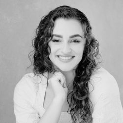 Smiling woman with long curly hair, wearing a light-coloured textured blouse, posing with her hand under her chin against a plain background (black and white portrait).