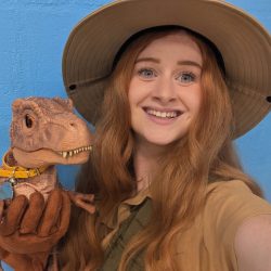 woman with long red hair weraing a safari hat and sqafari top holding a baby t rex